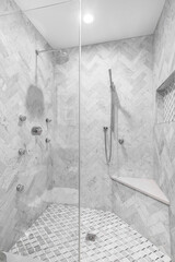 A luxury remodeled shower with marble tiles, a bench seat, and chrome faucets. The wall's are covered a herringbone tile pattern.