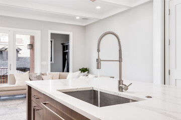 A kitchen sink with water running in a luxurious home looking out towards a living room area. The kitchen island features a white granite and stainless steel tub.