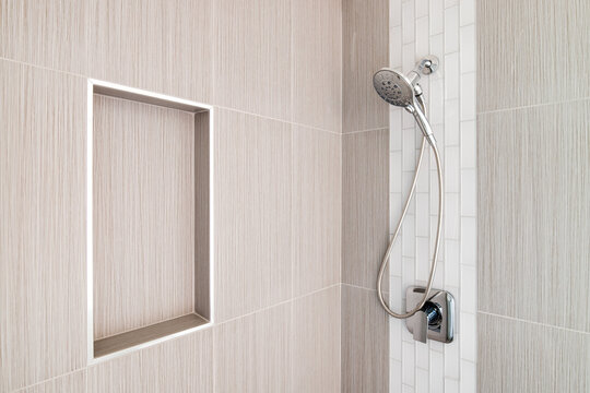 A detail shot of a beautifully tiled shower with brown large tiles, white subway tiles, and a shelf cut out for amenities. Chrome fixtures hang with a detachable shower head.