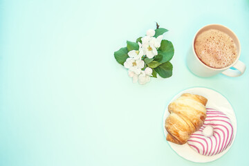 A composition of a cup of coffee with foam, a croissant and a spring apple blossomon a turquoise background . Good spring morning, flat lay.
