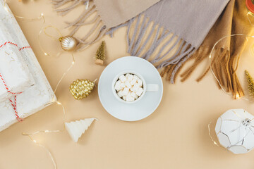 A cup of coffee with marshmallows, scarf, garlands, led lights on beige background. Women fashion winter clothes and accessories. New year and Christmas celebration mockup