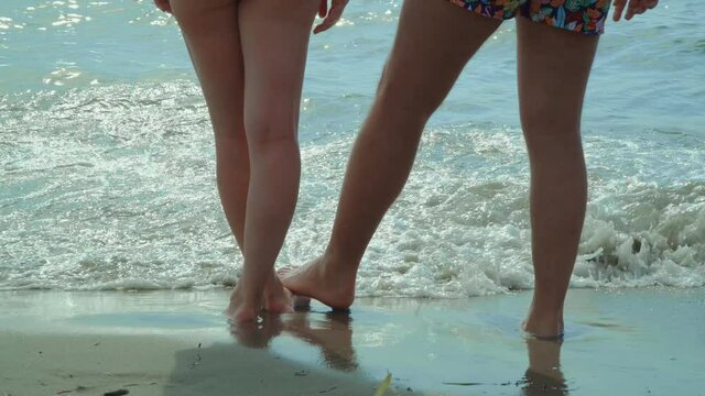 Couple's legs, feet standing and romantically touching on the sandy beach with the waves splashing on the sea shore