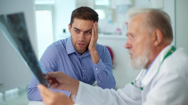 Professional mature male physician consult unhappy young man patient giving bad news explaining results of MRI image brain. Sad scared young man listening to bad news sitting in doctor office.