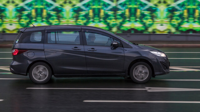 Mazda Premacy Third generation (CW) on wet city road. Dark gray compact minivan with man driving. Mazda5 auto in fast motion with blurred background.