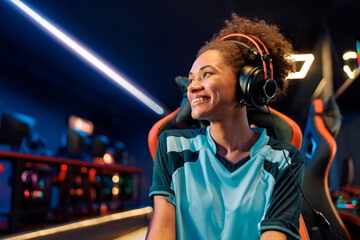 African female wearing wired headphones looking to side while sitting on gaming chair with happy...