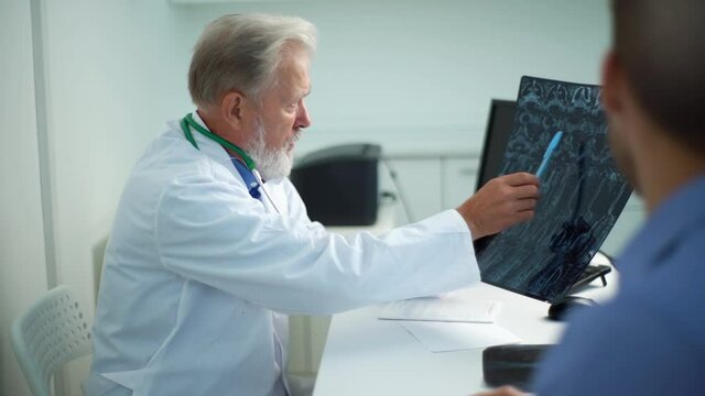 View from back of unrecognizable male patient listening mature adult doctor with MRI scan giving professional medical consultation for treatment illness, explaining causes of disease, slow motion.