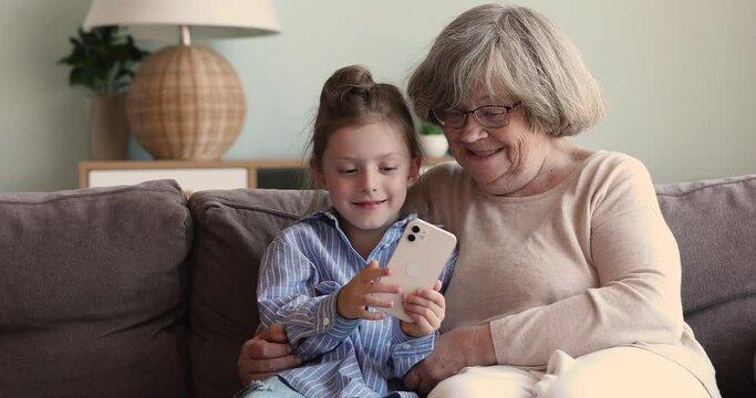 Cute little girl teach beloved older adult granny to make funny self picture on cell. Senior grandmother junior school age granddaughter rest on sofa enjoy taking selfies on phone holding victory sign