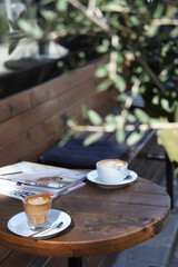 two coffee cups and a magazine are on the table. There is no people in photo. Olives leaves is blur in the photo.