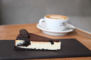 A slice of oreo cheesecake on the focus. One cup of coffee on the table. Dessert and coffee are the...