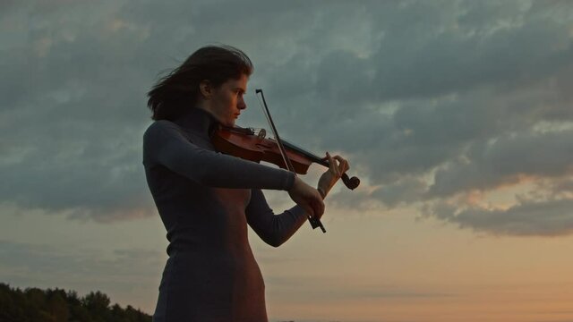 Woman musician is playing violin solo, standing outside, surrounded by nature, hair is fluttering on wind, harmony of nature and music art, Slow motion.