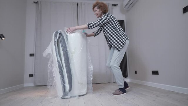 Happy adult woman unpacks new orthopedic mattress. Unpacking mattress with scissors pressed into bag. Moving to new apartment. New house without furniture. Elderly female opens mattress at home.