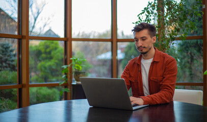 Fototapeta premium A man in a red shirt works at a laptop at a table during the day against the background of the garden.