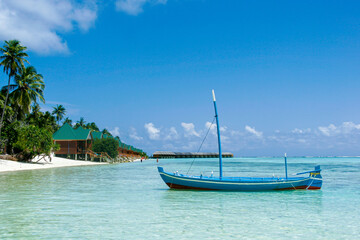 A traditional Maldivian boat called a dhoni moored in a sandy lagoon with beach bungalows in the background.
