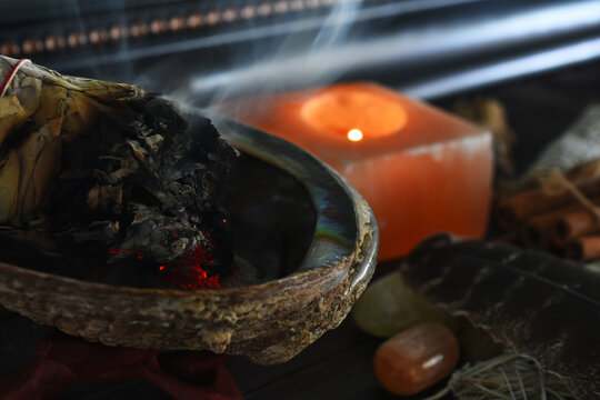 A close up image of a burning white sage smudge bundle and pink selenite candle. 