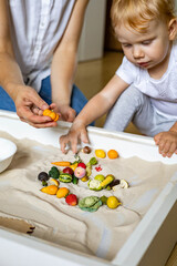 Obraz na płótnie Canvas Cheerful baby and mother playing tiny fruit vegetables wooden toys at home kinetic sandbox