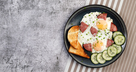 Fried eggs with green onions, sausages, toast and cucumber slices on a round plate on a dark grey background. Top view, flat lay