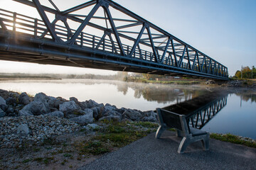 A steel girder bridge spans across a narrow river under blue skies and clouds. The deck of the...