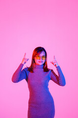 Studio shot Asian young female model in gray turtleneck sweater and orange lens sunglasses standing pointing index fingers to empty copy space presenting promoting product on pink light background