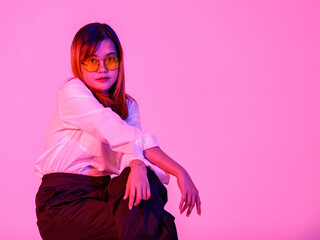 Portrait studio shot Asian young confident female designer entrepreneur model in casual fashionable wears with orange lens sunglasses sitting on chair posing look at camera on pink light background