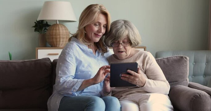 Caring older age woman grownup child of senior female retiree teach mom to use modern touchpad on sofa at living room. Happy old lady hold tablet pc talk to adult daughter show photo at social media