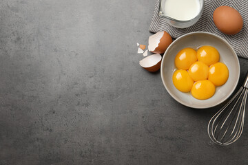 Bowl with raw egg yolks, whisk and jug of milk on grey table, flat lay. Space for text