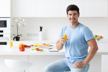 Handsome man in a blue T-shirt with a glass of orange juice in his hands in the kitchen at home