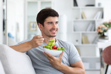 Handsome man eating fresh vegetable salad while sitting on the couch at home, healthy and vegan - obrazy, fototapety, plakaty