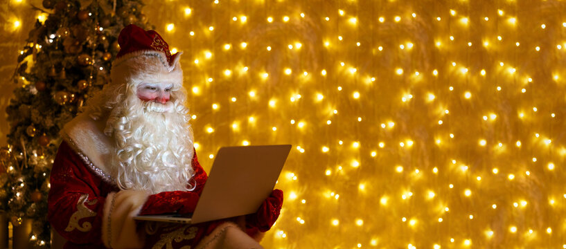 Christmas Online Greetings From Santa Claus. Santa Claus Using Laptop In Distance Christmas Call Kids Sitting Near Christmas Tree At Home. Happy New Year Covid. Against Background Of Wall With Garland