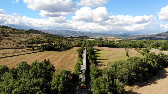 Aerial: old diesel train traveling along fields under summer blue sky with some clouds on a sunny day