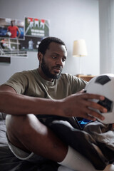 Contemporary African football player or trainer holding soccer ball while sitting on bed and preparing for training