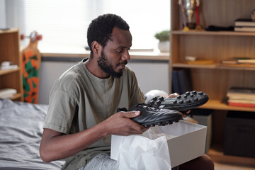 African man holding pair of new black trendy sports-shoes over while open box while unpacking sports footwear - obrazy, fototapety, plakaty