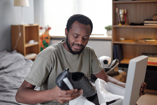 Young African sportsman unpacking box with new black sports-shoes while sitting on bed and preparing for football training - Powered by Adobe