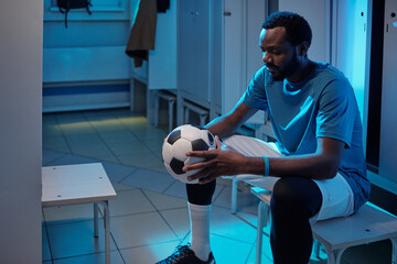 Young African sportsman looking at soccer ball in his hands while sitting by locker in changing-room at break