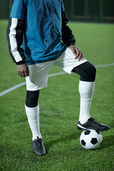 Young African football player in uniform keeping foot on soccer ball while standing on green lawn during game
