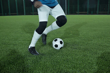 Legs of young active football player running after soccer ball while playing or training on green lawn at stadium