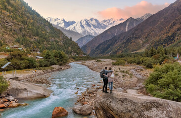 Tourist couple enjoy a vacation at the scenic Baspa river valley with view of Himalaya mountain range at Rakchham, Himachal Pradesh, India - obrazy, fototapety, plakaty