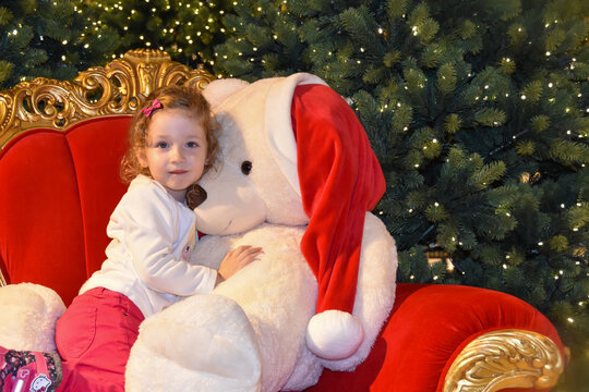 Weihnachtsstimmung. Ein kleines Mädchen kuschelt mit einem Teddybär auf einer roten Couch. Im Hintergrund Tannen.