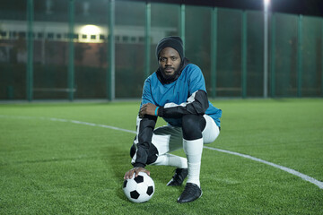 Young African footballer with soccer ball sitting on squats while having rest on football field...