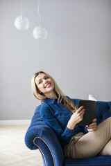 Portrait of a beautiful smiling blonde with a notebook in her hands in casual clothes in a blue cozy armchair.
