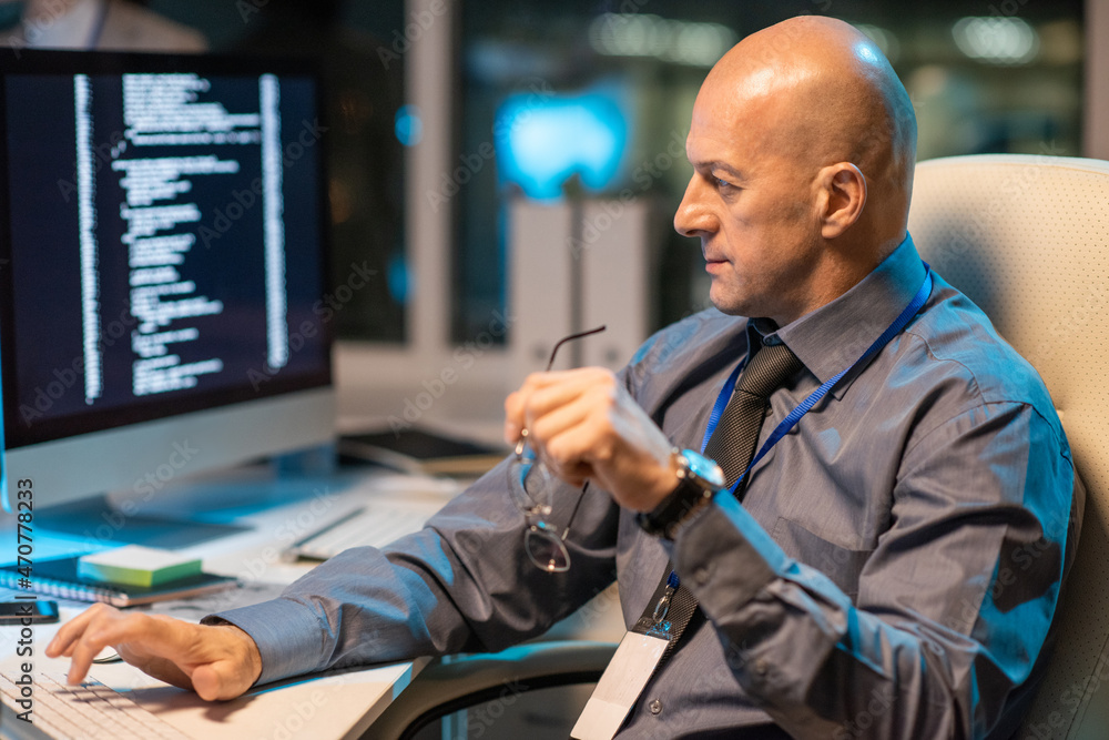Poster Mature bald it-engineer working in front of computer monitor while sitting in armchair by desk in openspace office