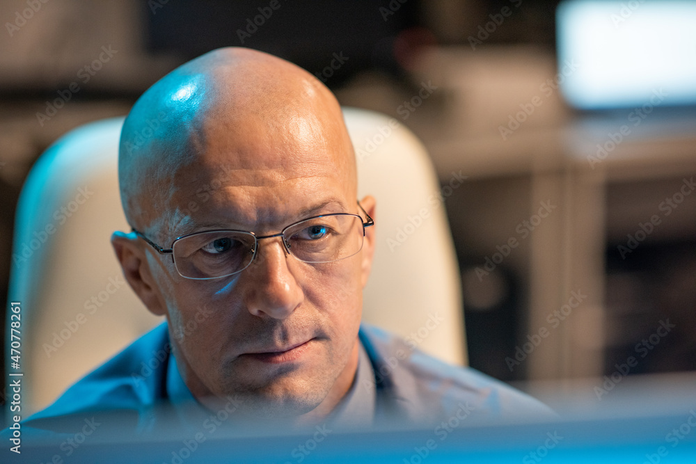 Poster experienced businessman in eyeglasses looking at data on computer screen while sitting by his workpl