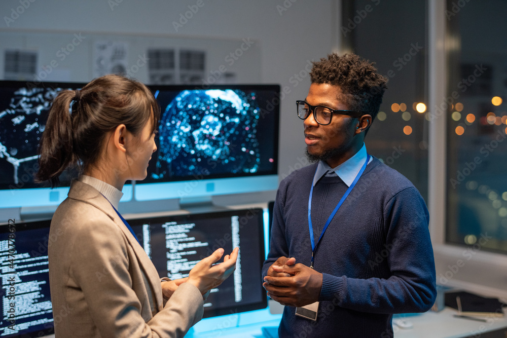 Poster two young multiracial colleagues having discussion against workplace with computer monitors in large