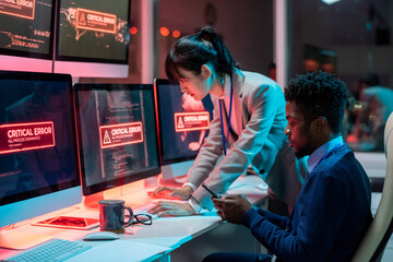 Two young intercultural programmers trying to solve technical problem in front of computers with critical error on screens