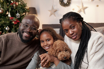 Happy young African couple and their daughter with cute pet sitting in front of camera and looking at you