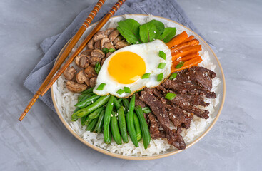 Korean Bibimbap cuisine made of boiled rice topped with beef slices, green beans, carrot, mushrooms and fried egg with yolk served on plate with wooden chopsticks on gray concrete table with towel
