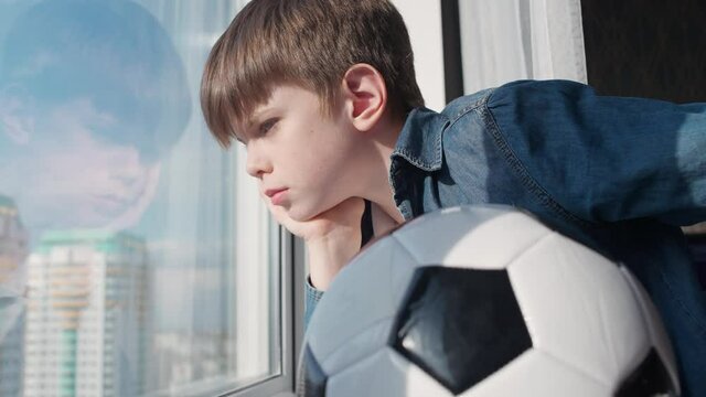 Caucasian boy on a home quarantine looking out the window, a bored boy holds a soccer ball, self-isolation in the context of the coronavirus pandemic, reflection in glass, childhood during lockdown.