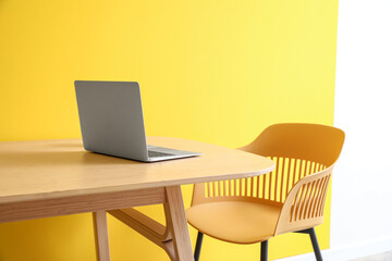 Modern laptop on wooden table and chair near color wall