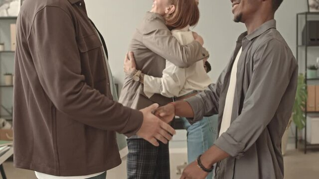 Tilting up of four diverse young colleagues greeting, hugging and shaking hands in office after long break, talking and smiling in morning