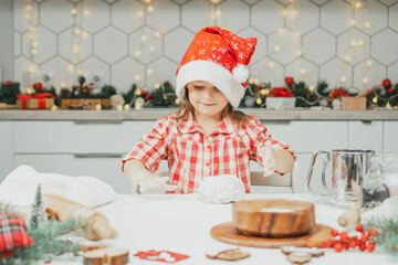 Little girl 3 years old in red Christmas cap and checkered shirt prepares dough for gingerbread cookies in kitchen