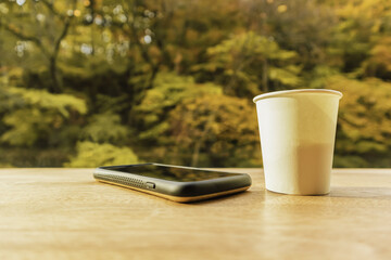 white paper hot coffee cup on wooden table with blurred autumn tree background outside the window.